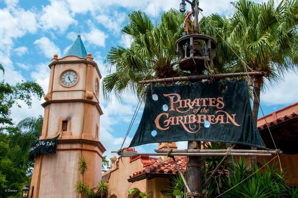 A Pirates of the Caribbean attraction entrance sign and clock tower at Disney theme park beneath blue skies and palm trees.