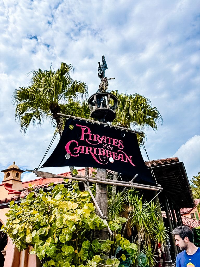 Pirates of the Caribbean attraction entrance sign with palm trees and cloudy blue sky at Disney theme park.