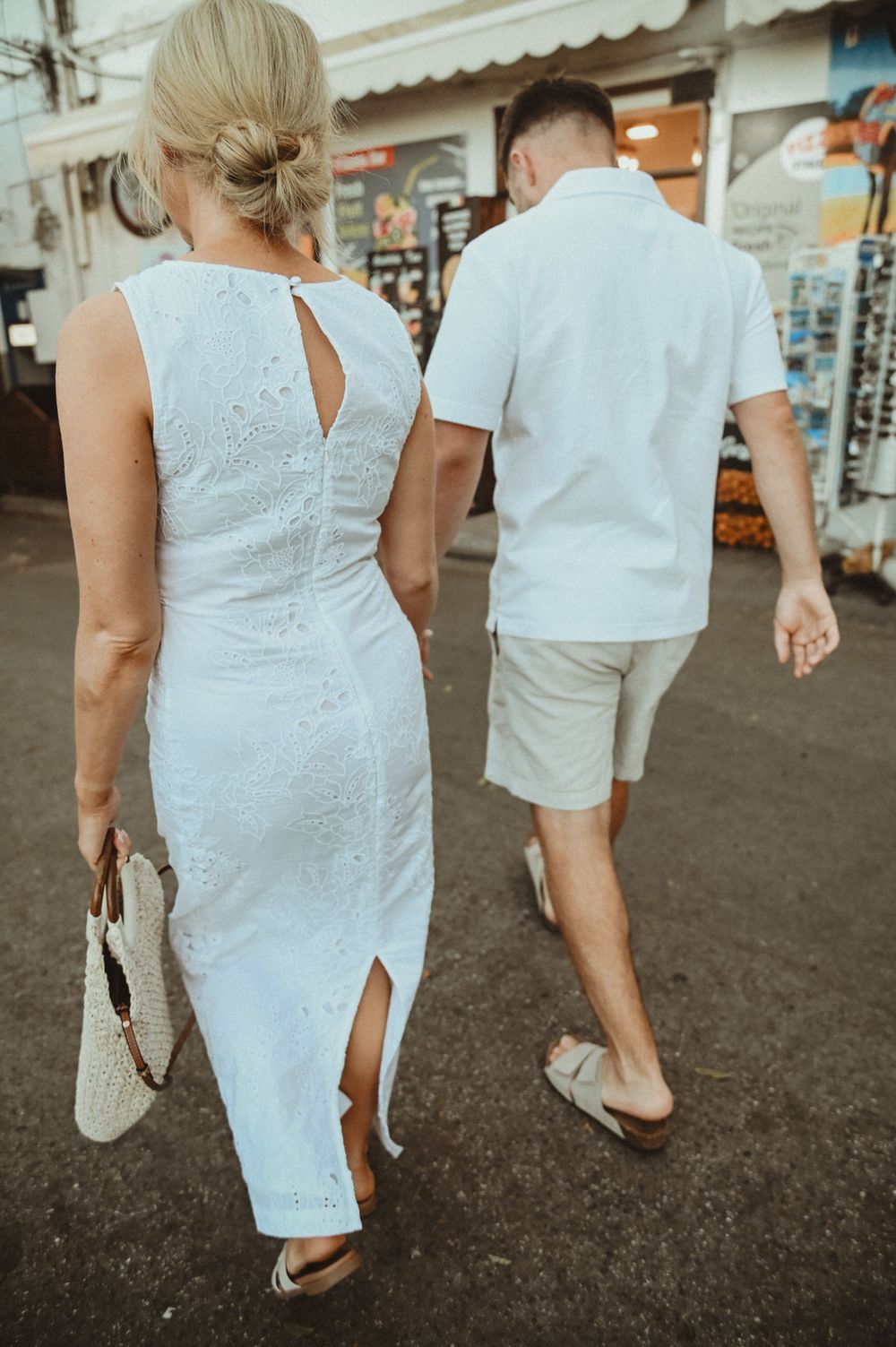 A couple walks down a street wearing coordinated white summer outfits with the woman in a lace dress carrying a woven bag.