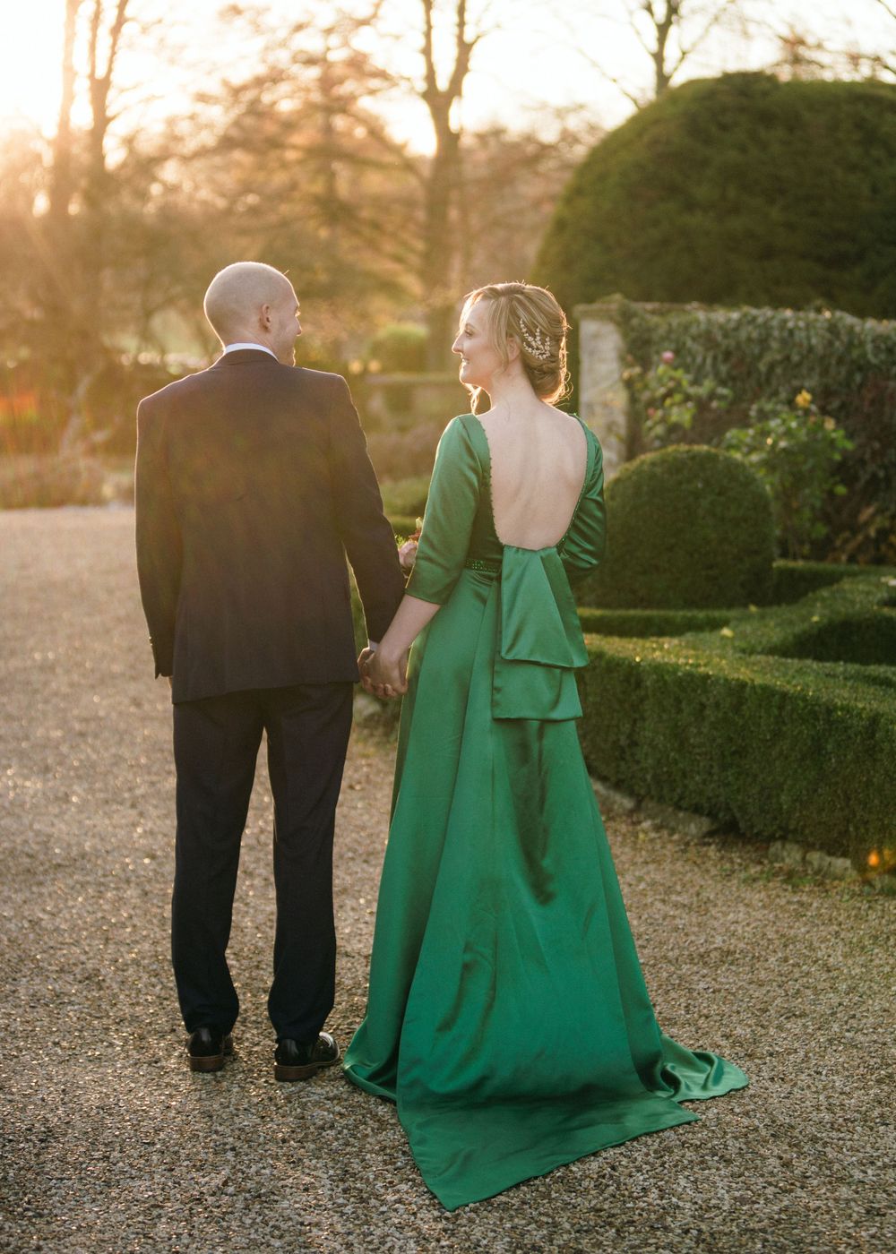 A couple walks along a garden path at sunset in Saraguay House, Nova Scotia, woman wearing an elegant emerald green backless gown.