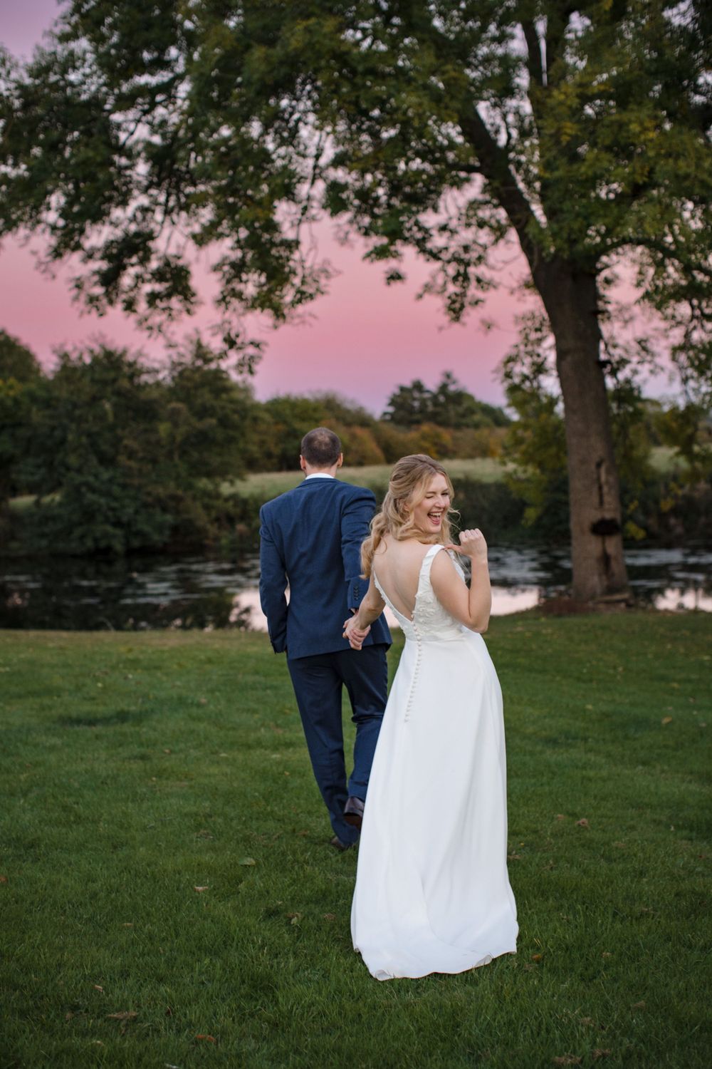 A couple in wedding attire stands by a lake at sunset with pink skies and trees framing the romantic scene Oakfield Golf and Country Club