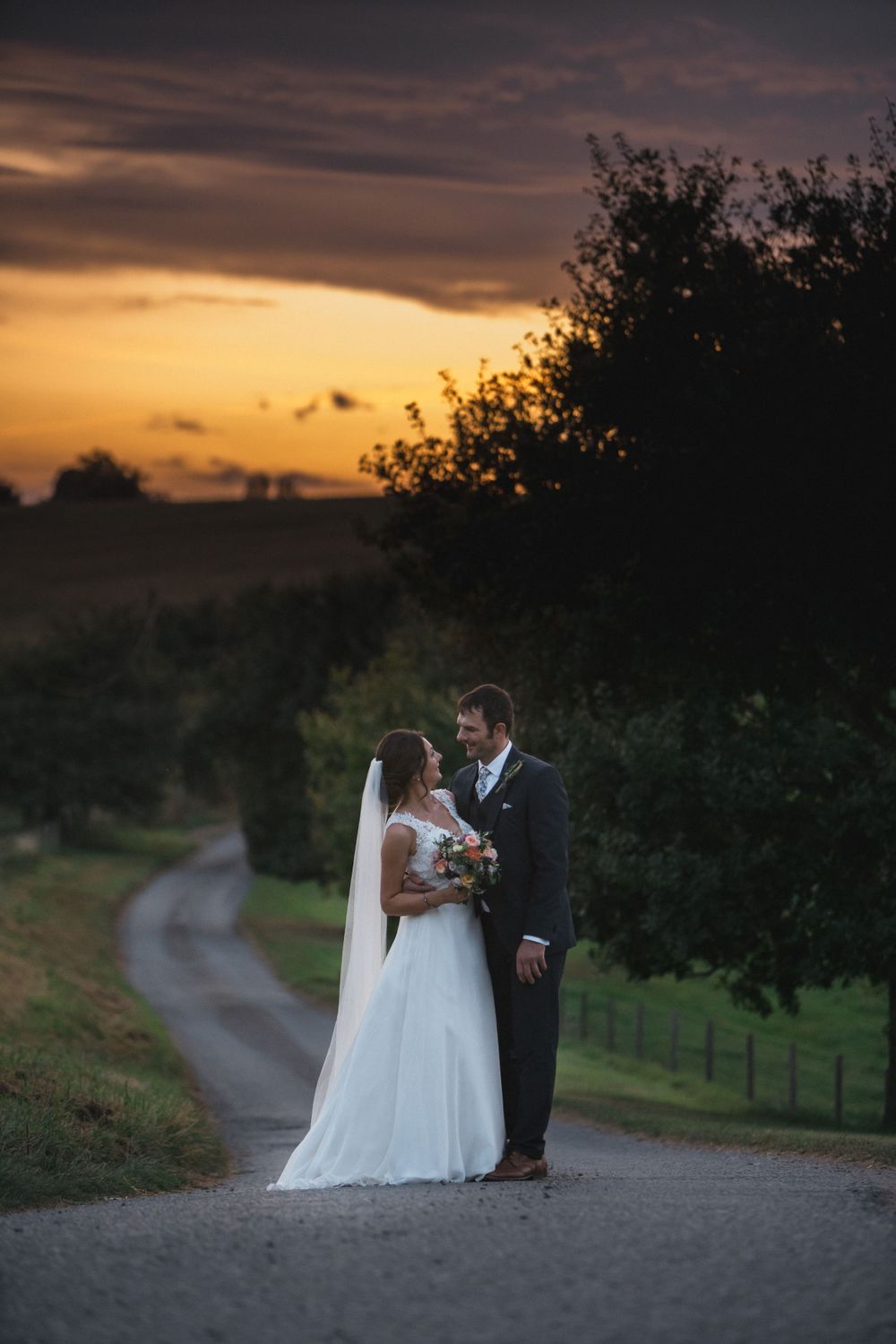 A beautiful sunset wedding photo of a couple standing on a winding country road with trees silhouetted against the orange sky Saraguay House