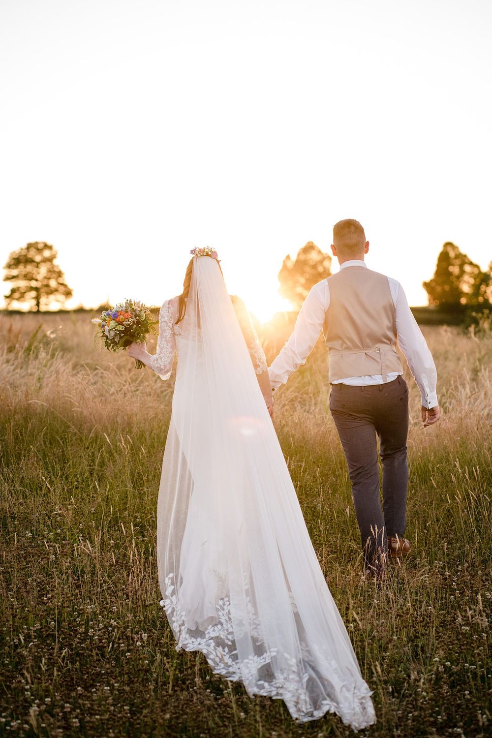 A couple walk together through a sunlit meadow at golden hour with her lace veil trailing behind Lightfoot and Wolfville, Nova Scotia.