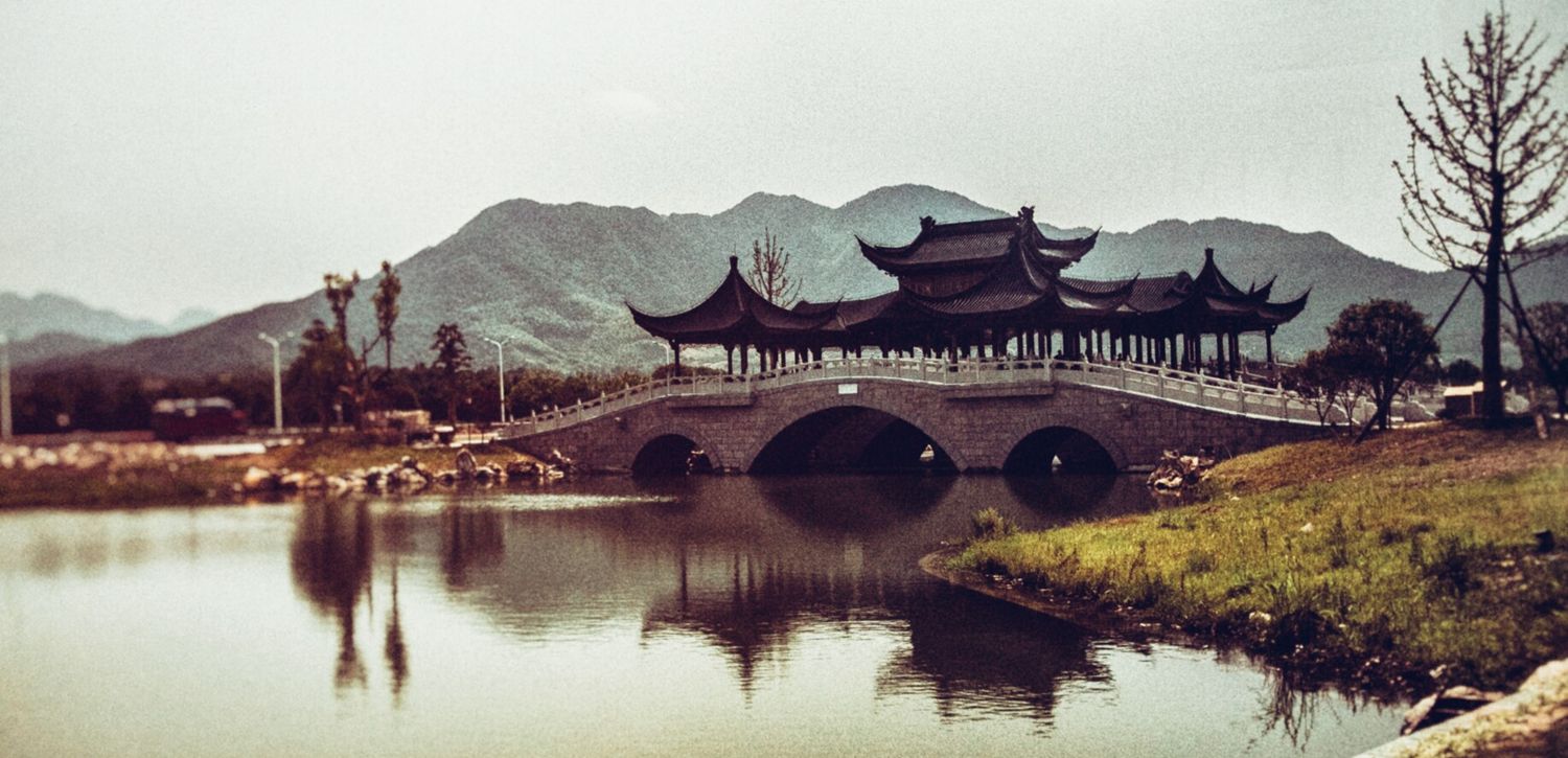 Traditional Asian bridge and pagoda buildings reflect in a serene lake with mountains in the background.