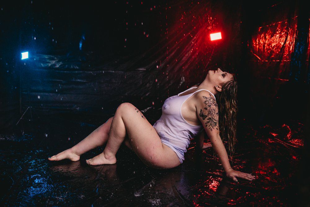 Artistic portrait with dramatic red and blue lighting shows figure in white dress reclining on wet ground during nighttime shoot.