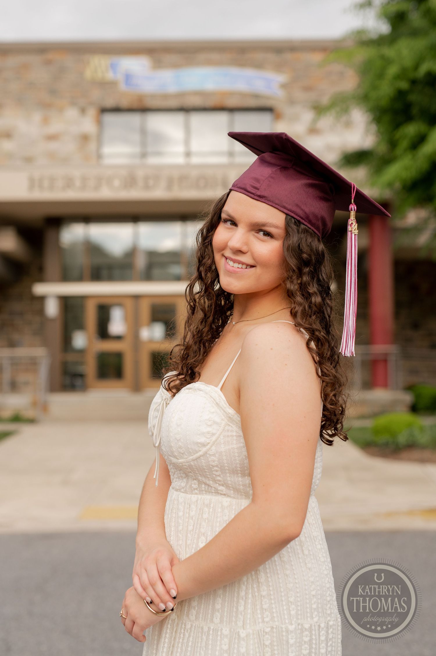 Graduate in maroon cap and white dress poses in front of brick school building.