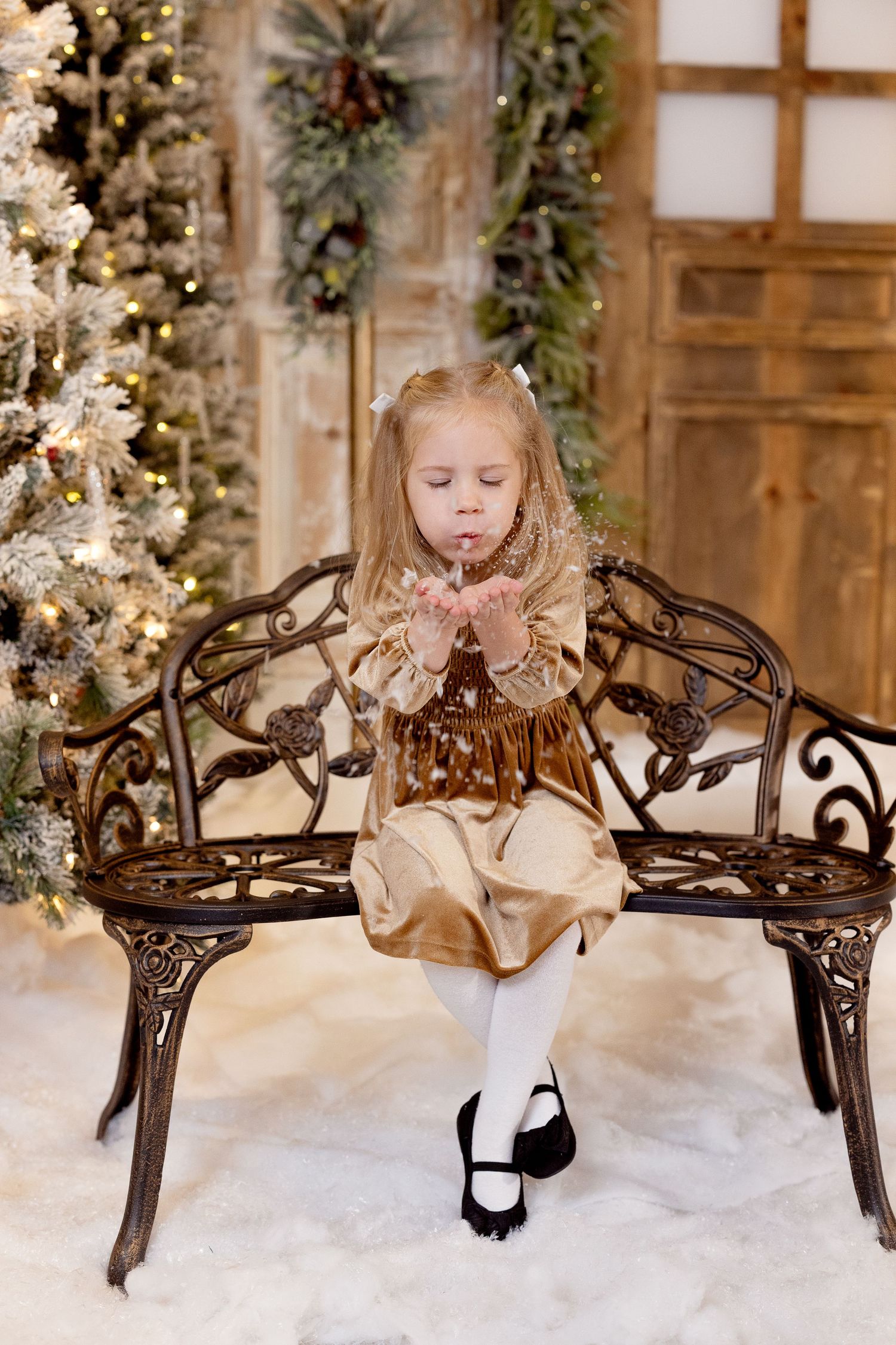 A child in a holiday dress sits on a decorative metal bench near a flocked Christmas tree and festive holiday decor.