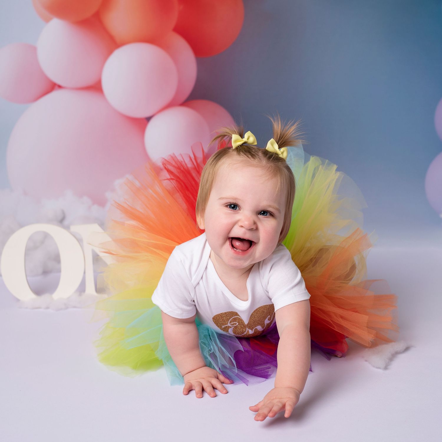 Baby wearing colorful rainbow tutu crawling on floor with pink balloon decorations in background.