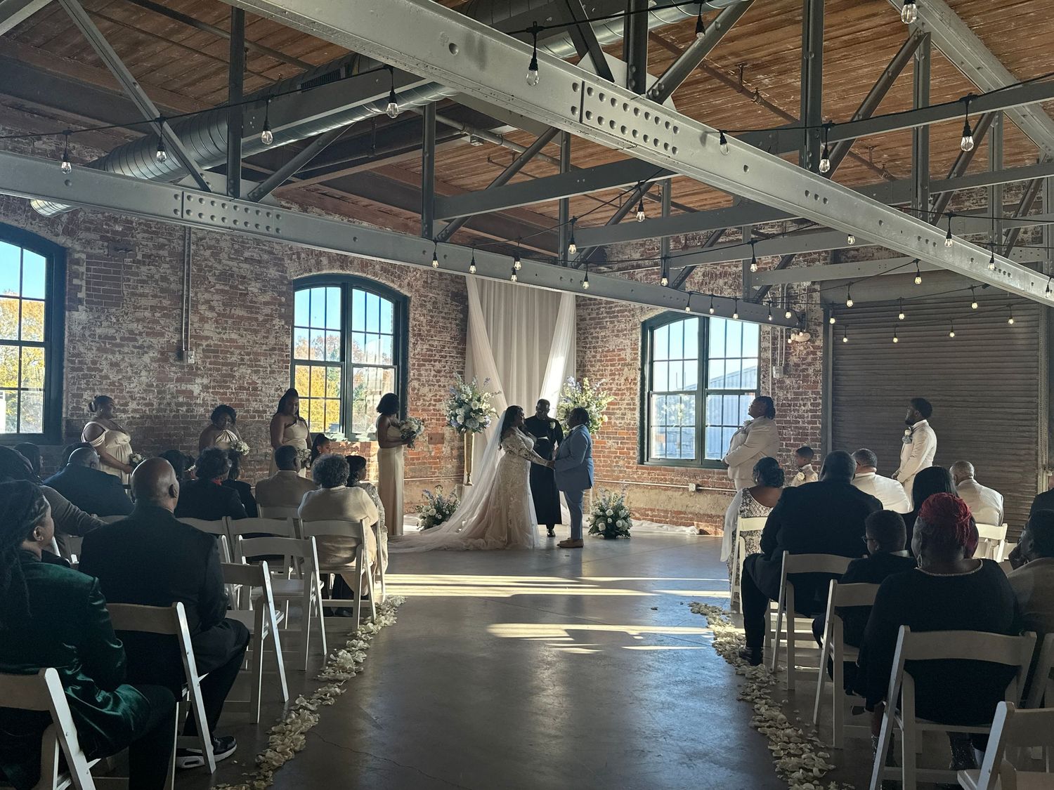 Wedding ceremony in an industrial venue with exposed brick walls and string lights overhead.
