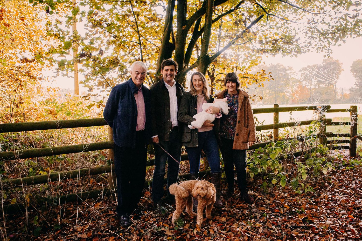 Extended family gathers for a group photo on a wooden fence during fall with their dog.