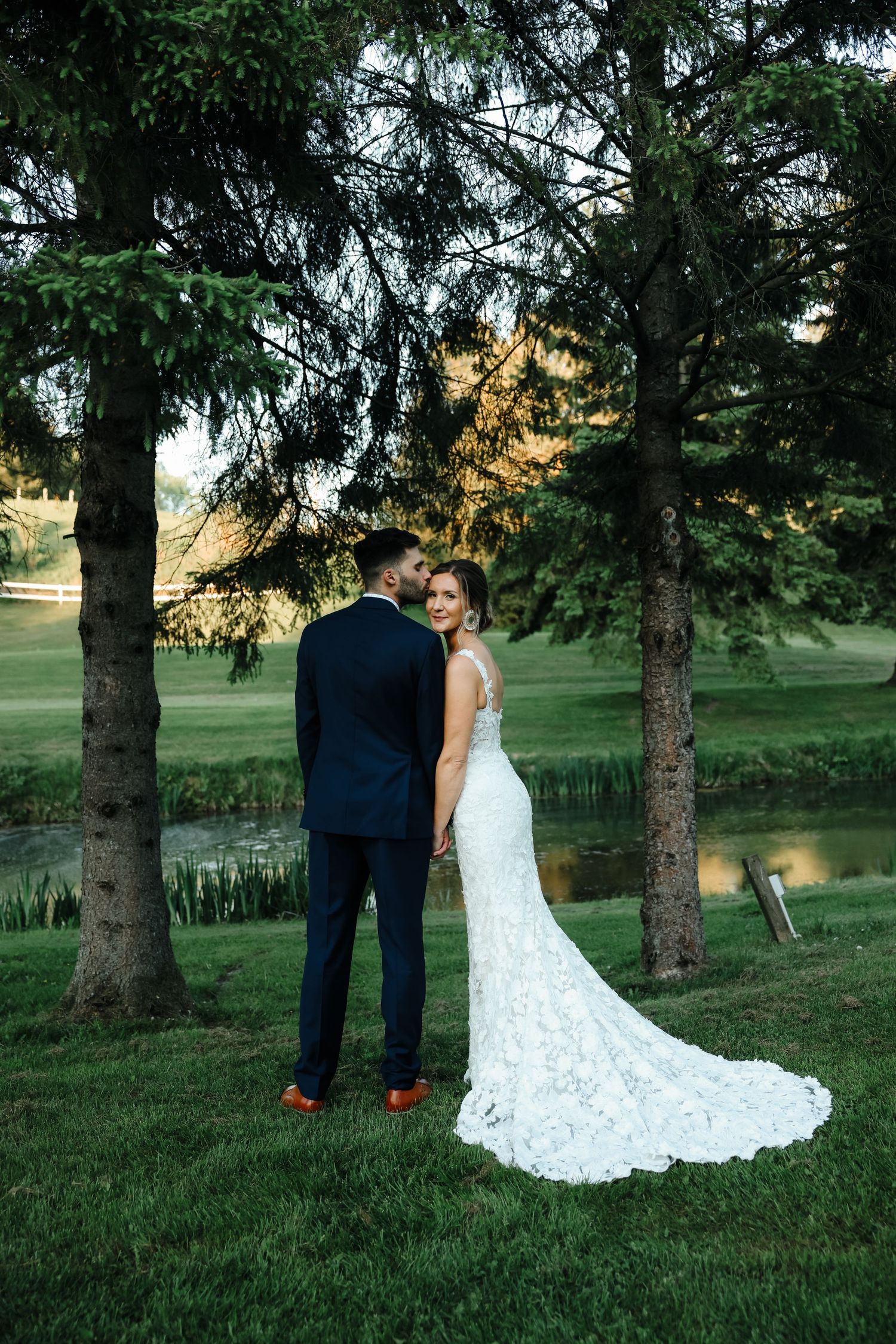 Wedding couple in formal attire standing among evergreen trees at sunset.