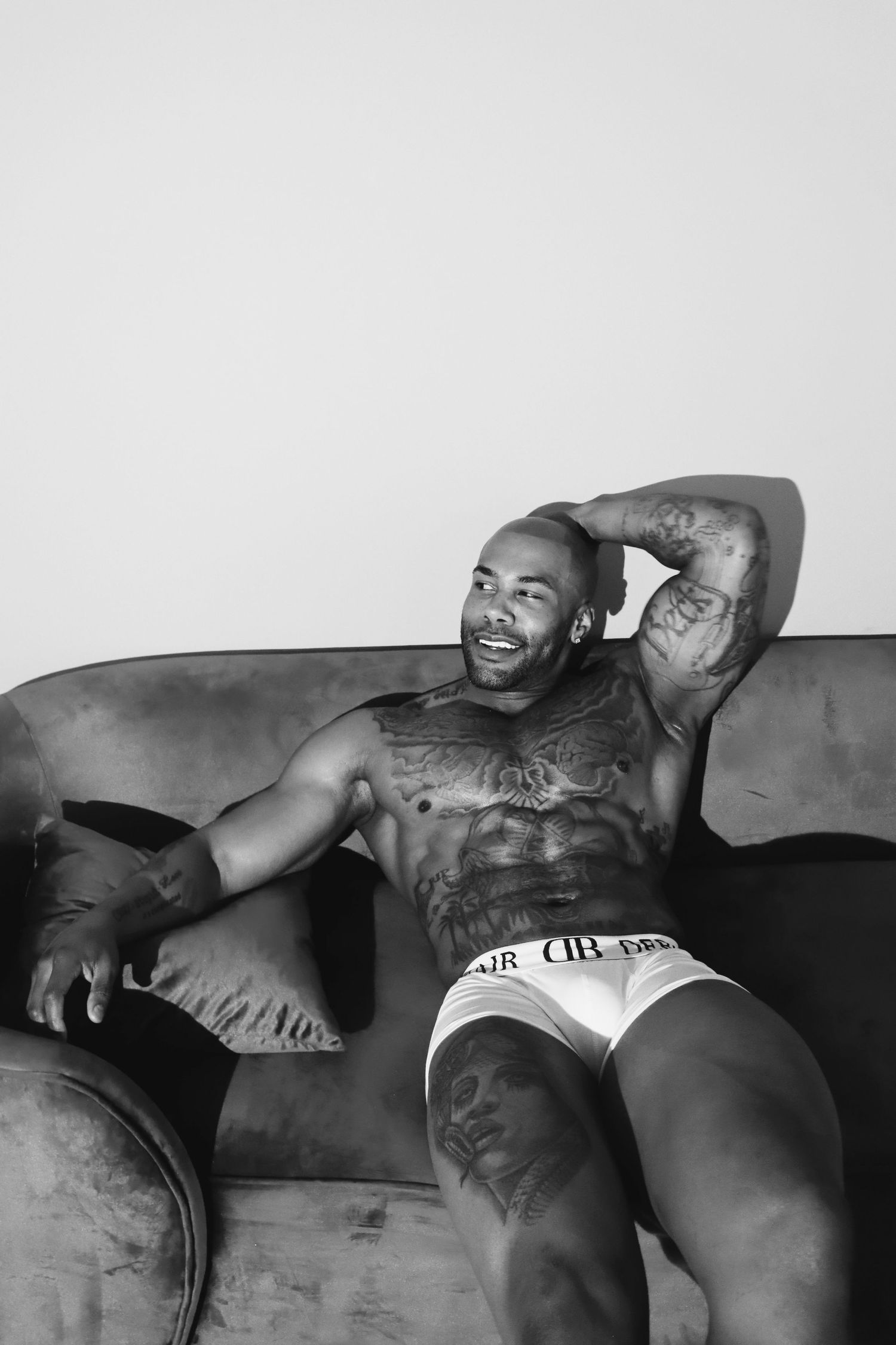 Black and white portrait of someone relaxing shirtless on a couch wearing white underwear and displaying tattoos.