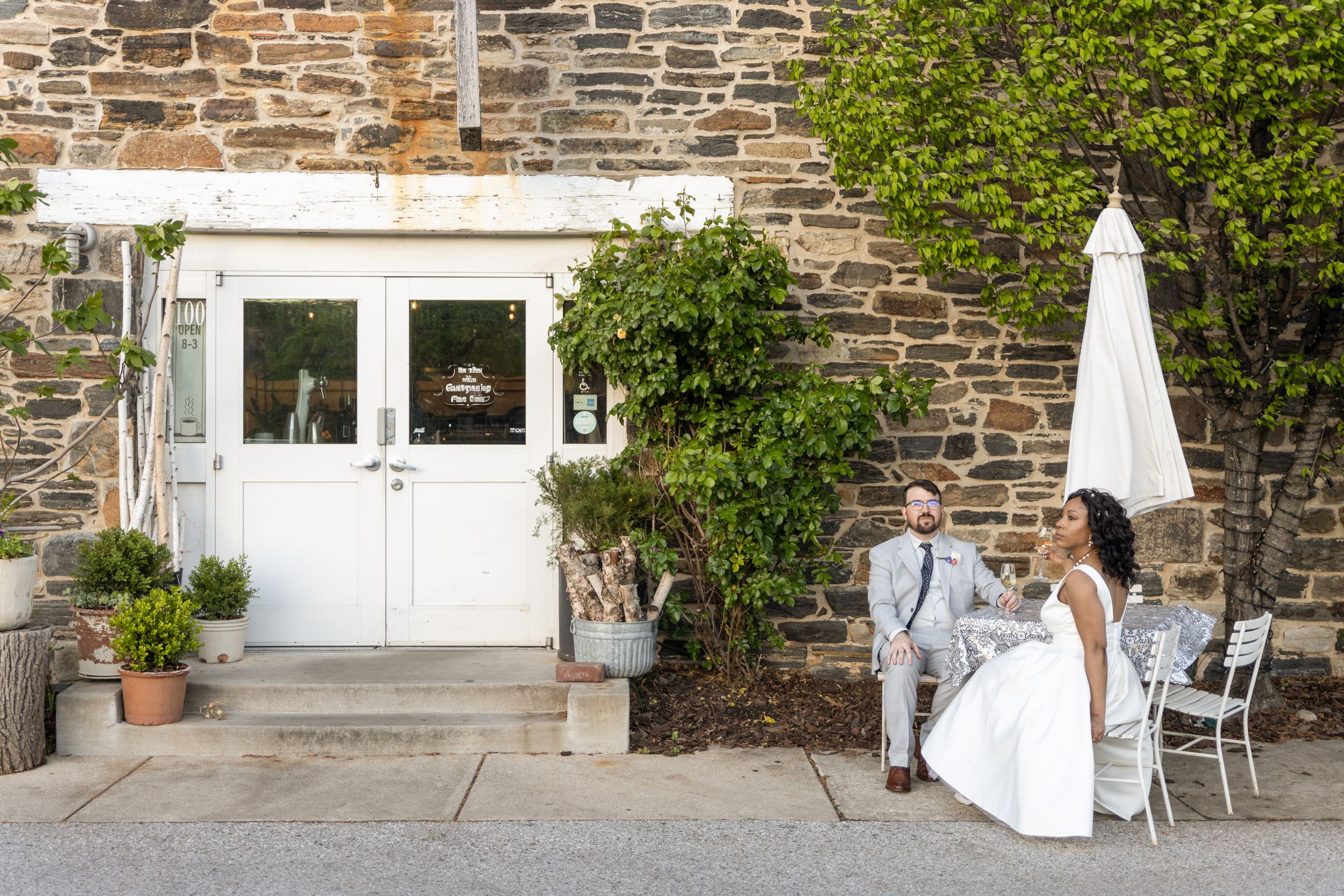 A couple in formal attire stands outside rustic stone building entrance with white doors and climbing greenery.