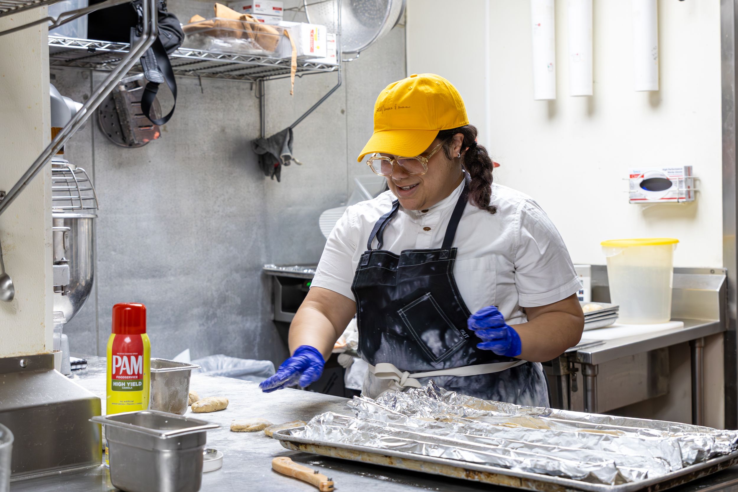 A kitchen worker in protective gear and yellow cap preparing food in an industrial kitchen setting.