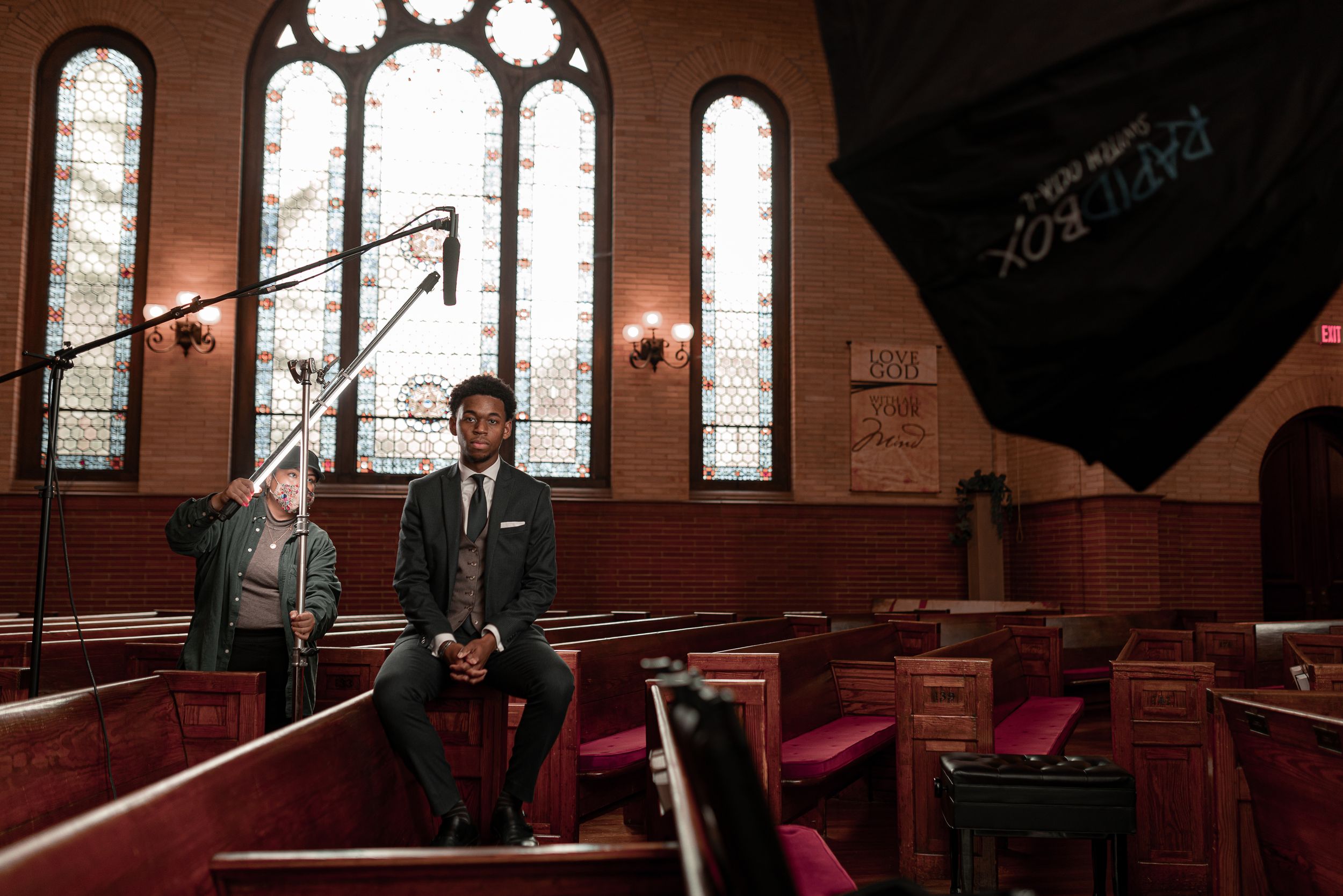 A person in a suit sits alone on a wooden church pew with stained glass windows in the background.