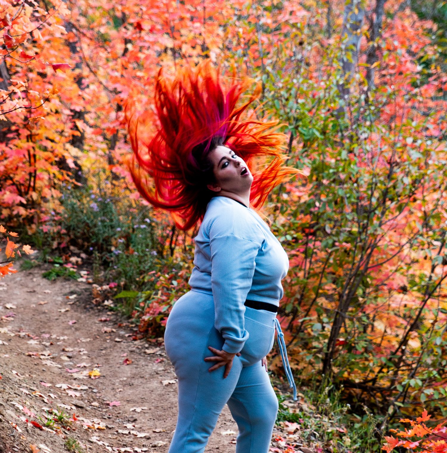 Woman with vibrant red hair tossing it dramatically amid colorful autumn foliage on a trail.