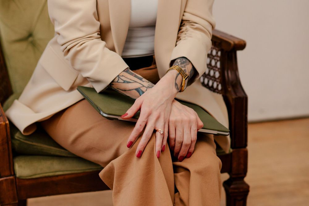 Person wearing a beige blazer and pants sits in an ornate wooden chair with green upholstery, showing hands and wristwatch.