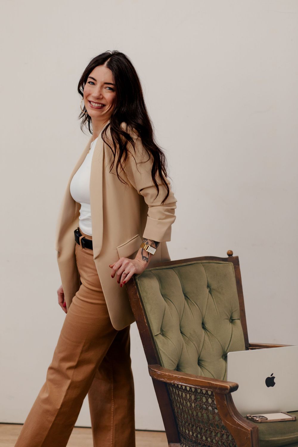 Professional wearing beige blazer and brown trousers poses beside vintage green tufted chair with laptop.