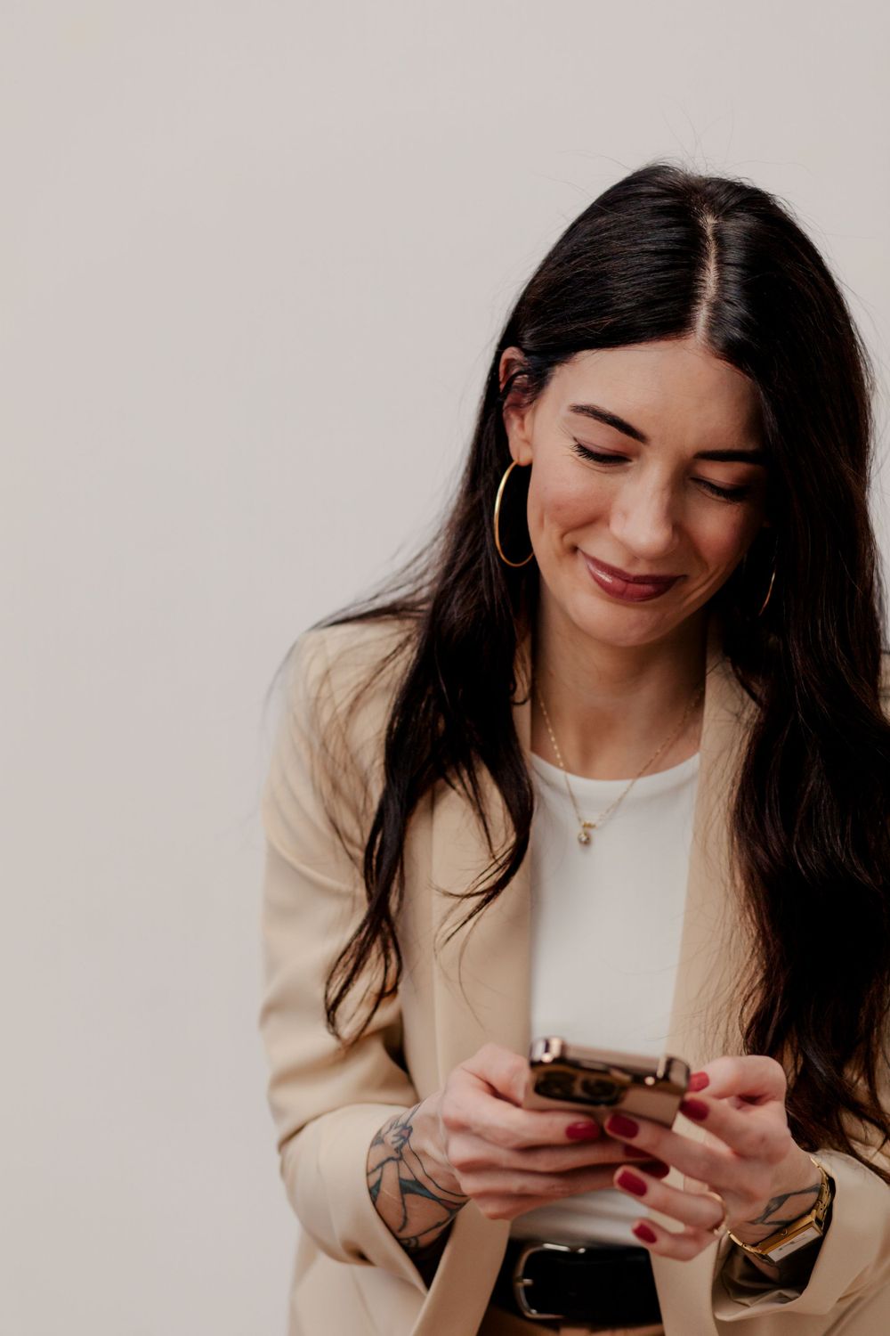 Person with long dark hair in beige blazer and white top checks phone while smiling.