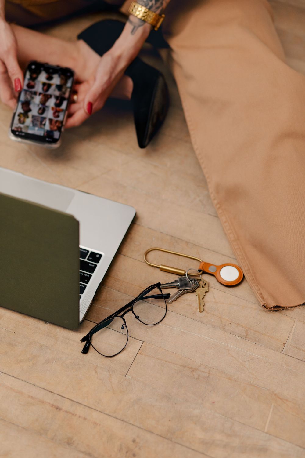 Flatlay workspace setup with laptop, glasses, AirTag, and phone on wooden surface.