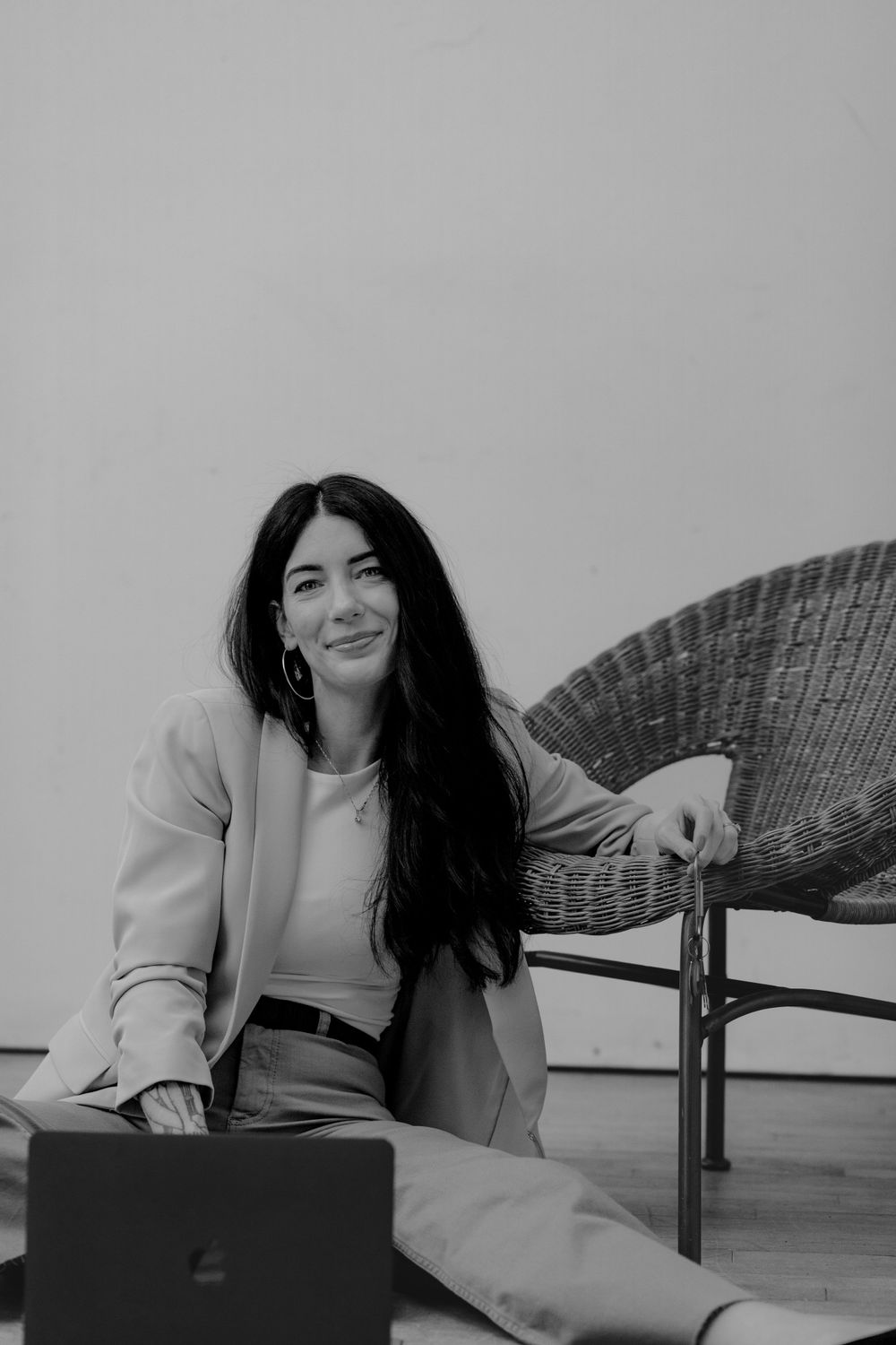 Black and white image showing someone sitting on floor with laptop next to a wicker chair wearing professional attire.