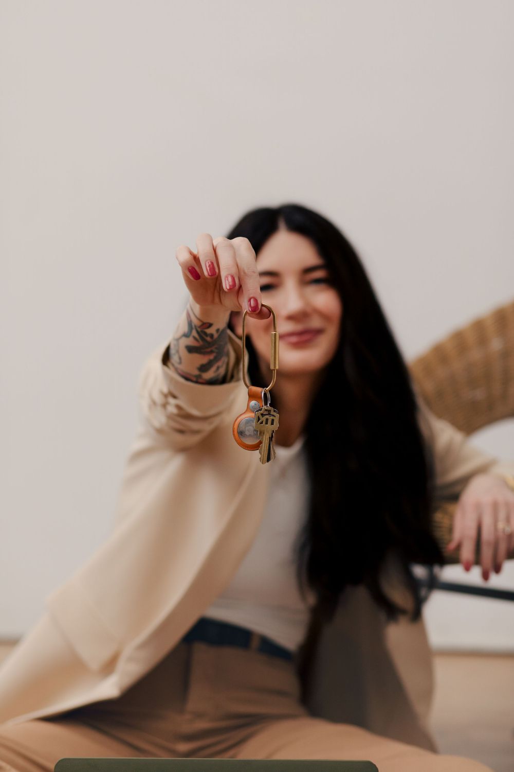Person in beige blazer and brown pants holds up keys while sitting, with long dark hair and joyful expression.