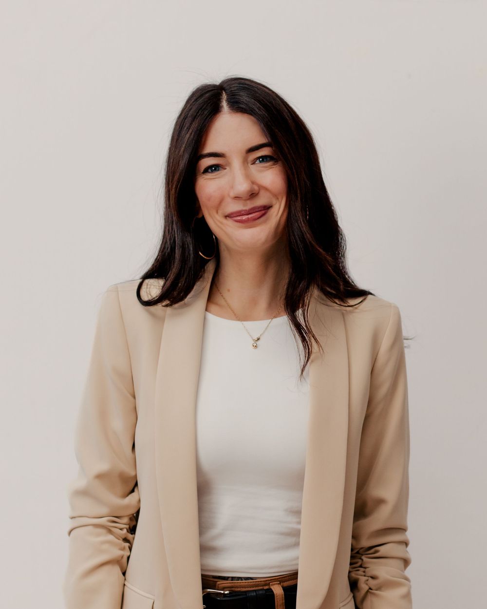 Professional portrait of someone with dark hair wearing a beige blazer and white top against a light background.