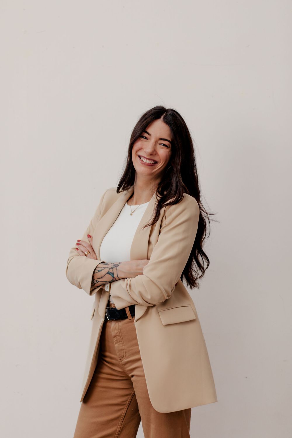 Professional wearing beige blazer and brown pants poses with crossed arms against neutral wall with bright smile.