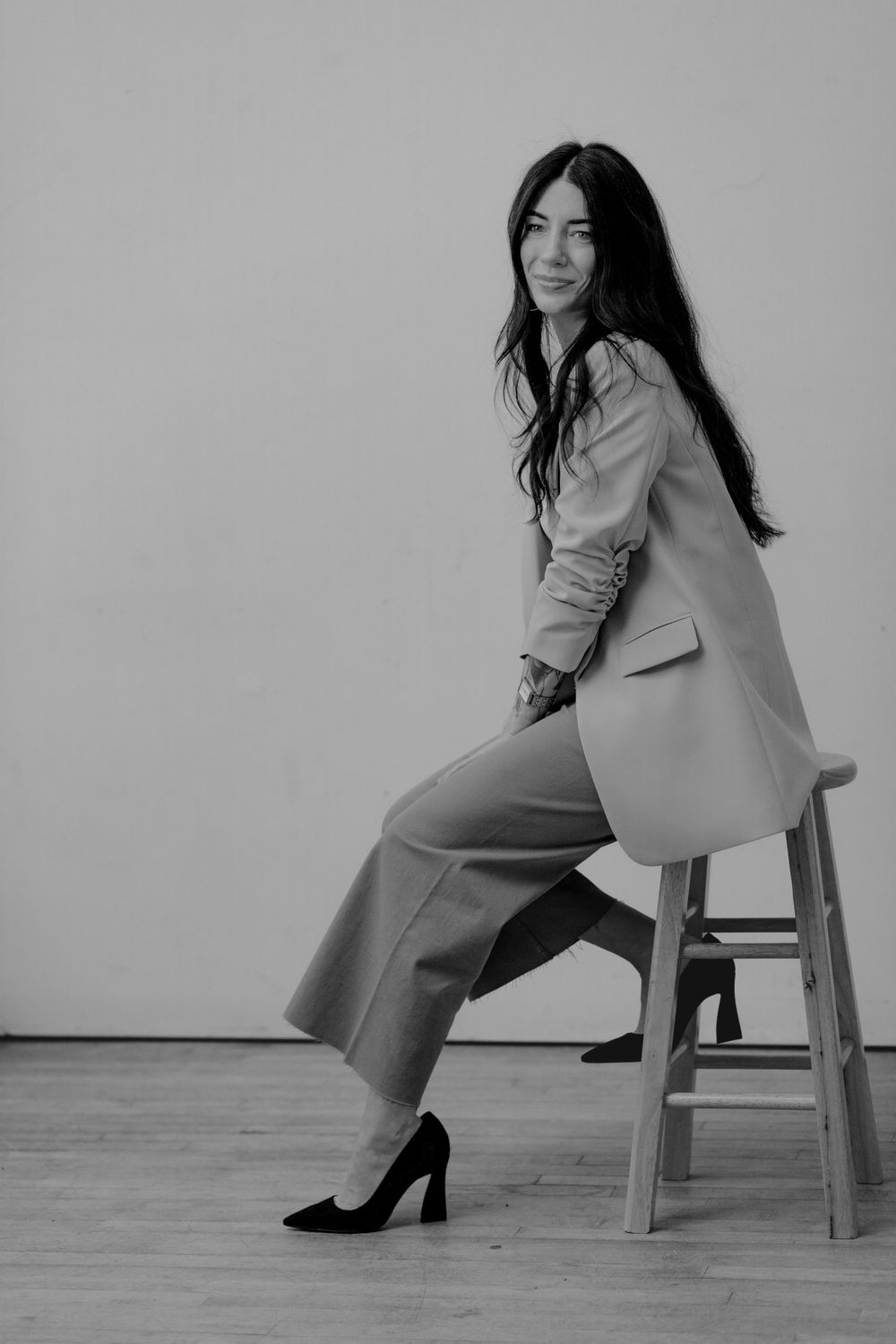 Black and white portrait of person in business attire sitting sideways on wooden stool against plain wall.