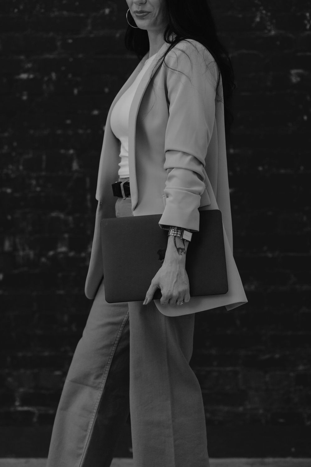 Professional wearing light blazer and carrying laptop case against dark brick wall in black and white.