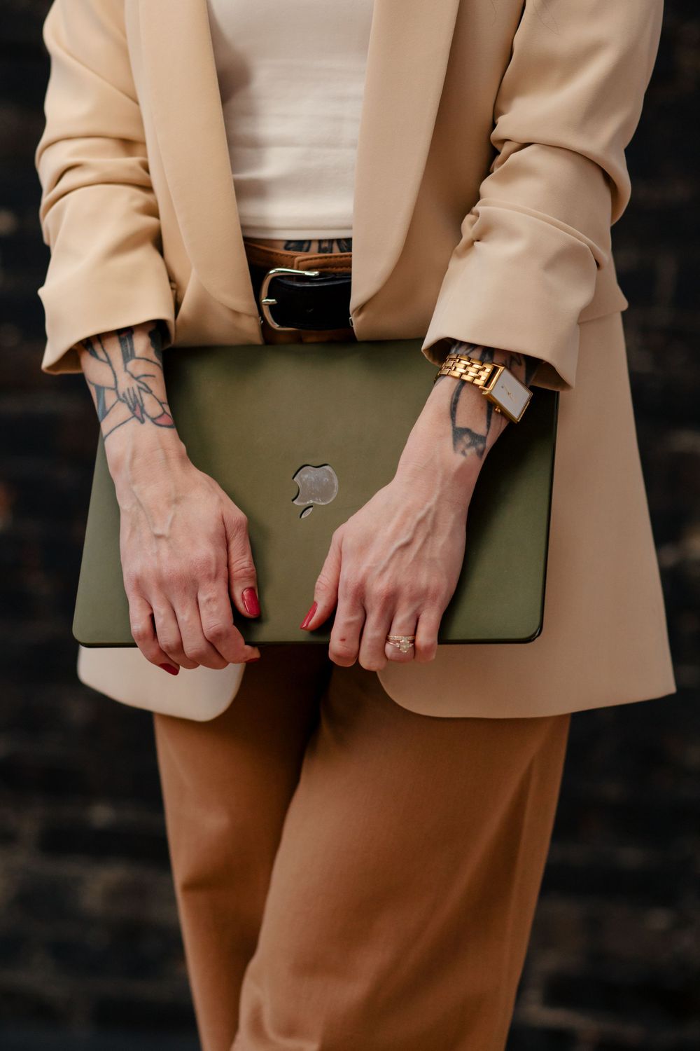 Hands holding an olive green laptop case against a beige blazer and brown pants with visible tattoos and red nails.