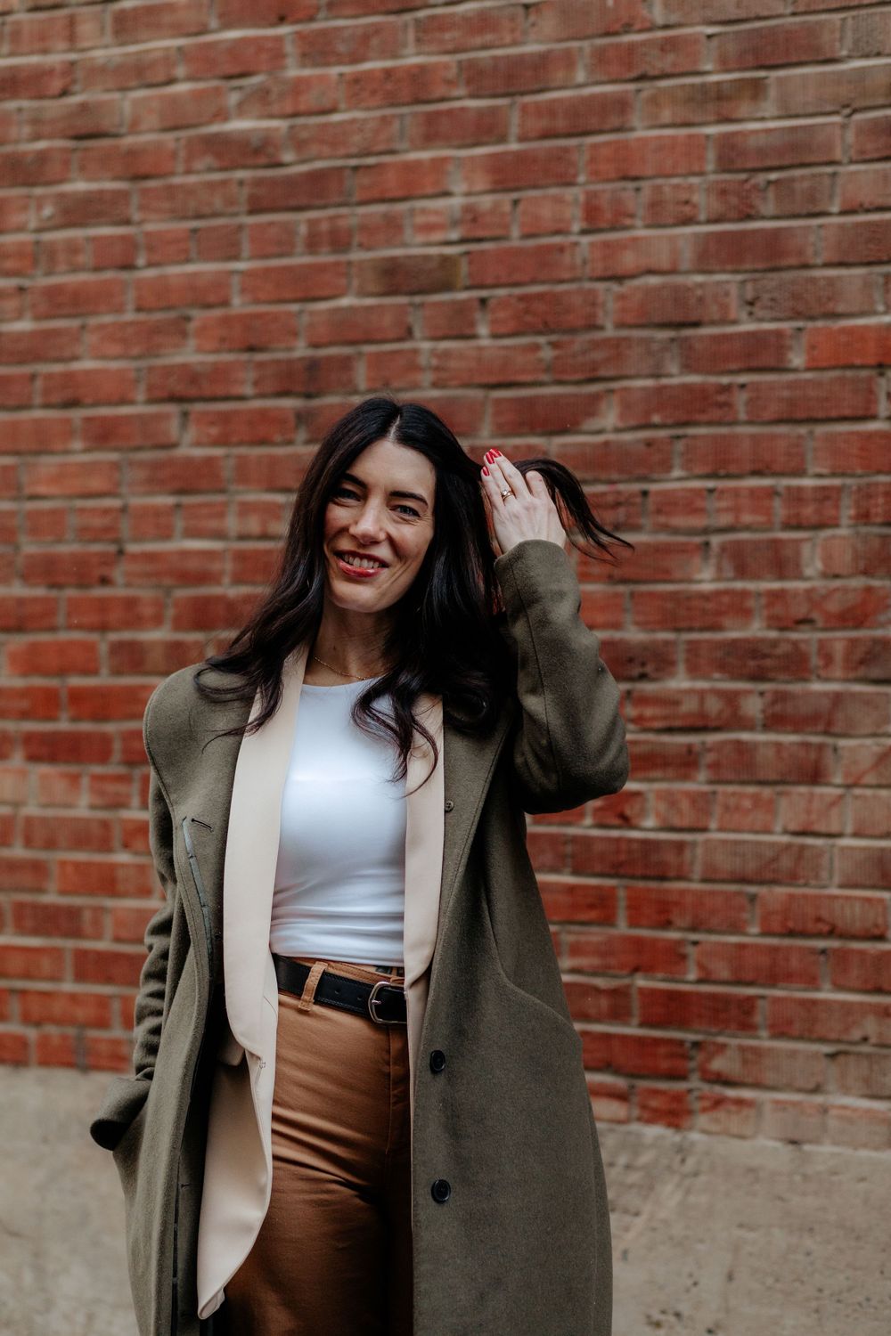 Person in olive coat and white top standing against red brick wall.