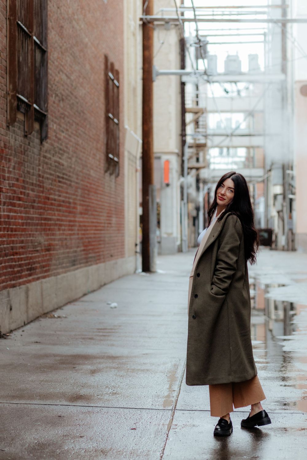 A figure in a long olive coat poses playfully in an urban alley with brick walls on a cloudy day.