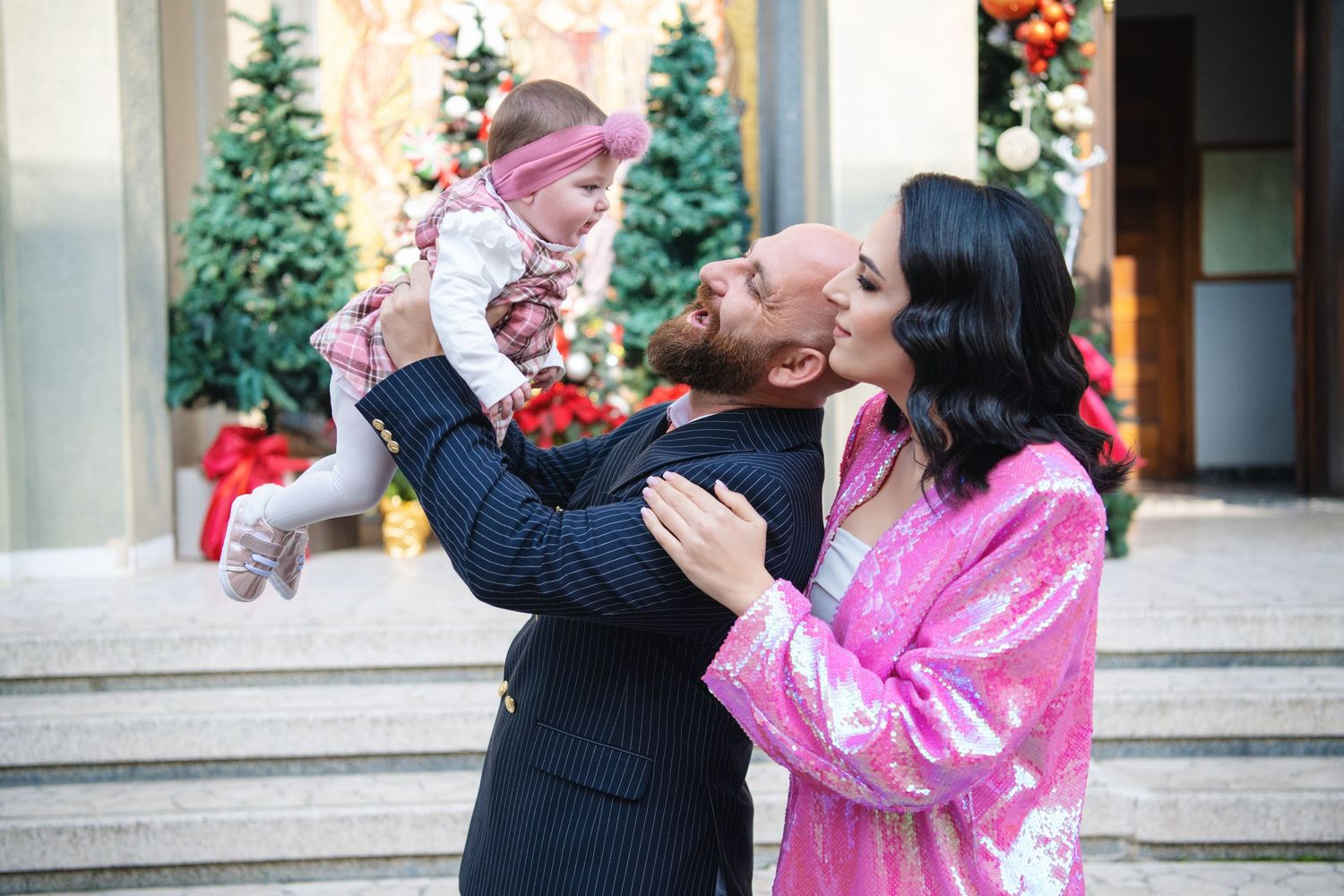 A couple in pink and black attire share a joyful moment with their baby outdoors during winter.
