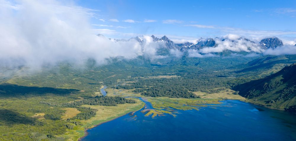 Panoramic aerial view of snow-capped mountains, lush valleys, and pristine lakes in the wilderness under blue skies.