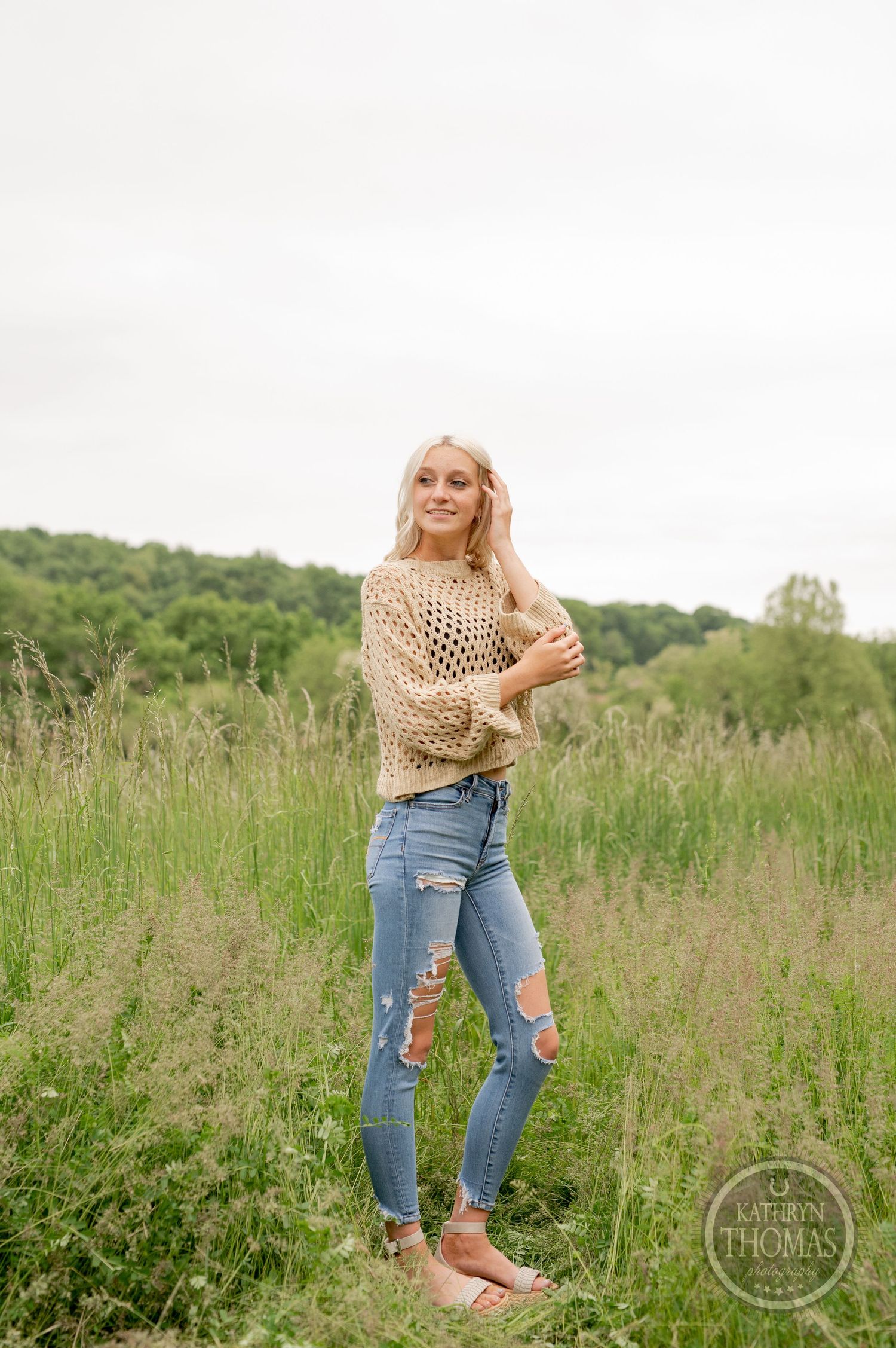 Natural outdoor fashion shot featuring a beige mesh sweater and ripped jeans against green countryside.