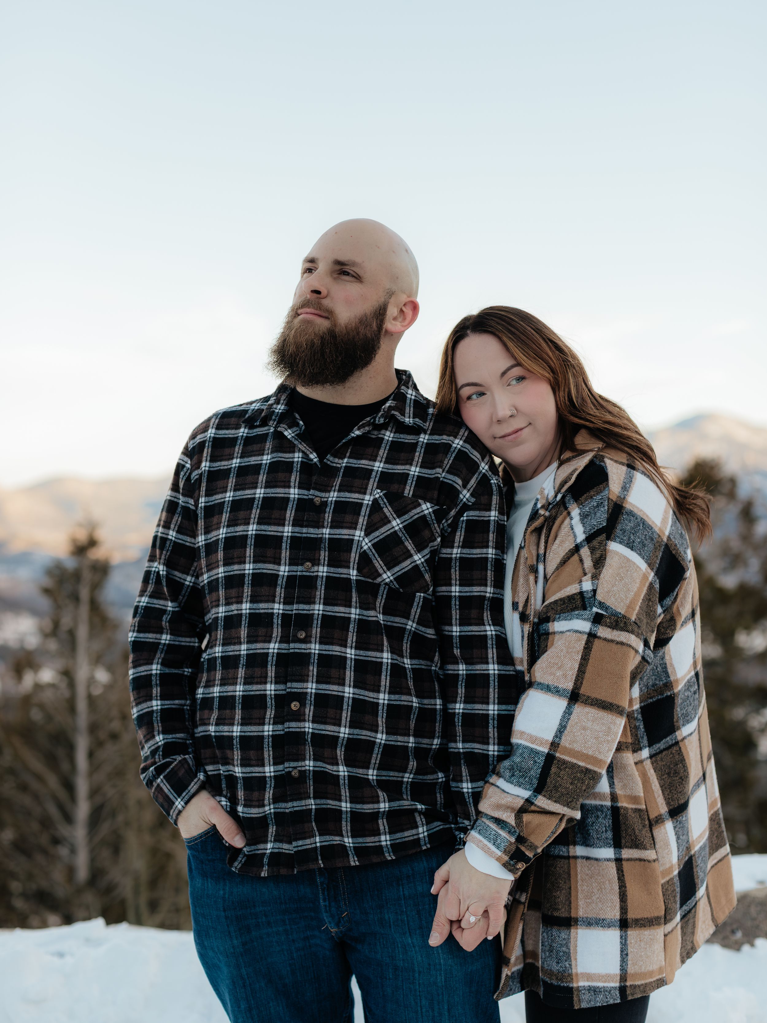 A couple in plaid and tan jackets embrace while standing in snowy winter scenery during an outdoor photoshoot.