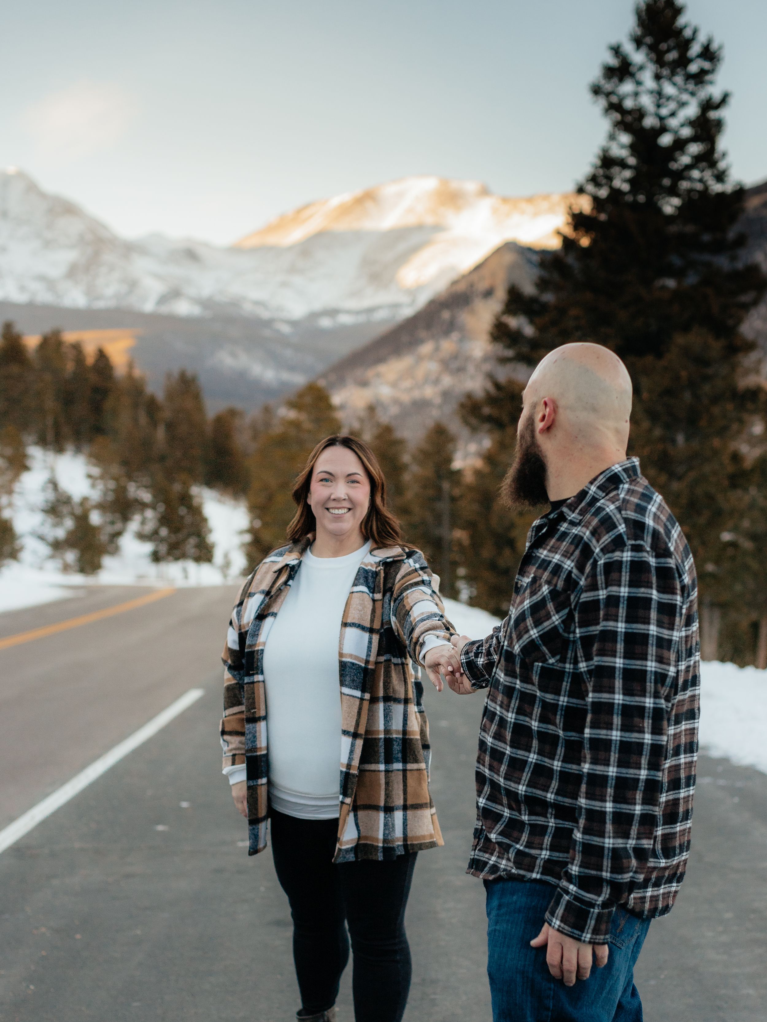 A couple shares a joyful moment together on a snowy mountain road with snow-capped peaks in the background.
