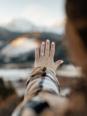 Hand wearing an engagement ring held up against a blurry mountain landscape background in warm lighting.