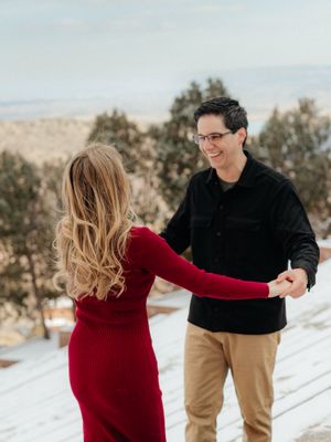 A couple in a snowy winter landscape, with her wearing a red dress and him in a black jacket and khaki pants.