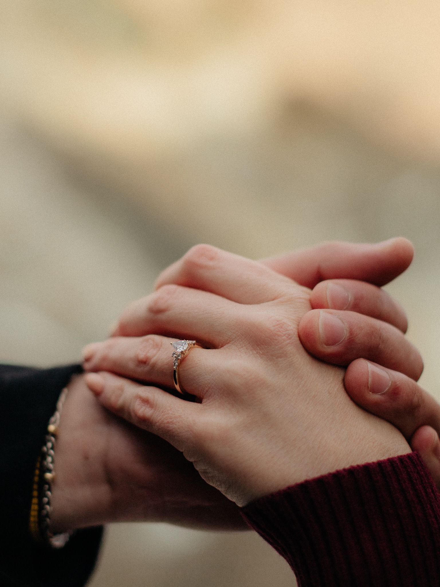 Two hands clasped together intimately showing a delicate engagement ring against a blurred warm background.