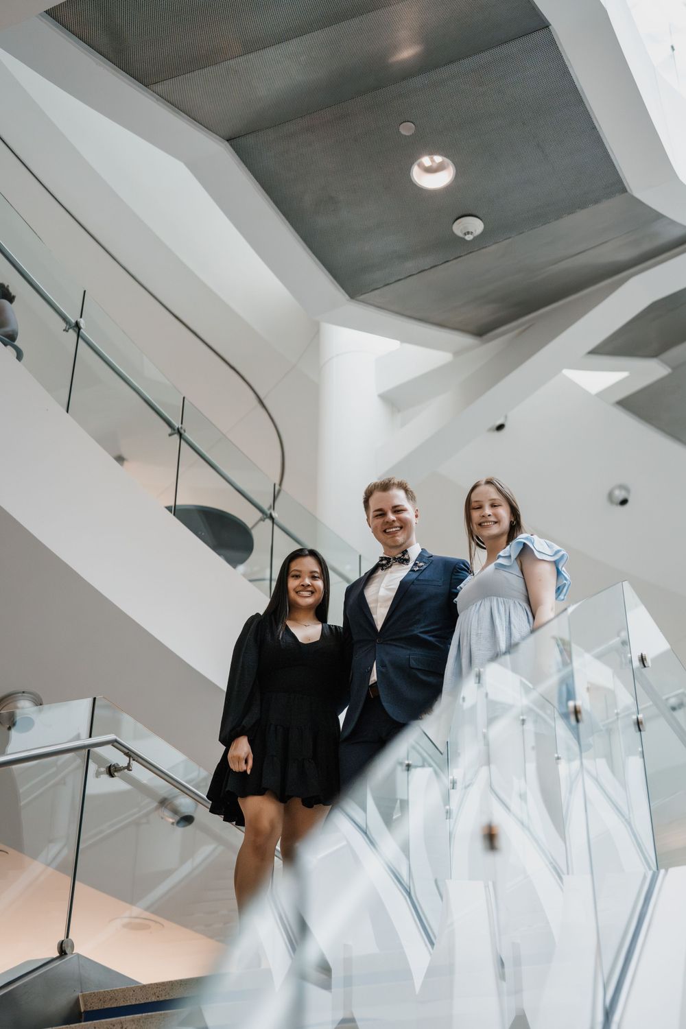 Three people posing on modern white stairs with glass railings in an architectural setting.
