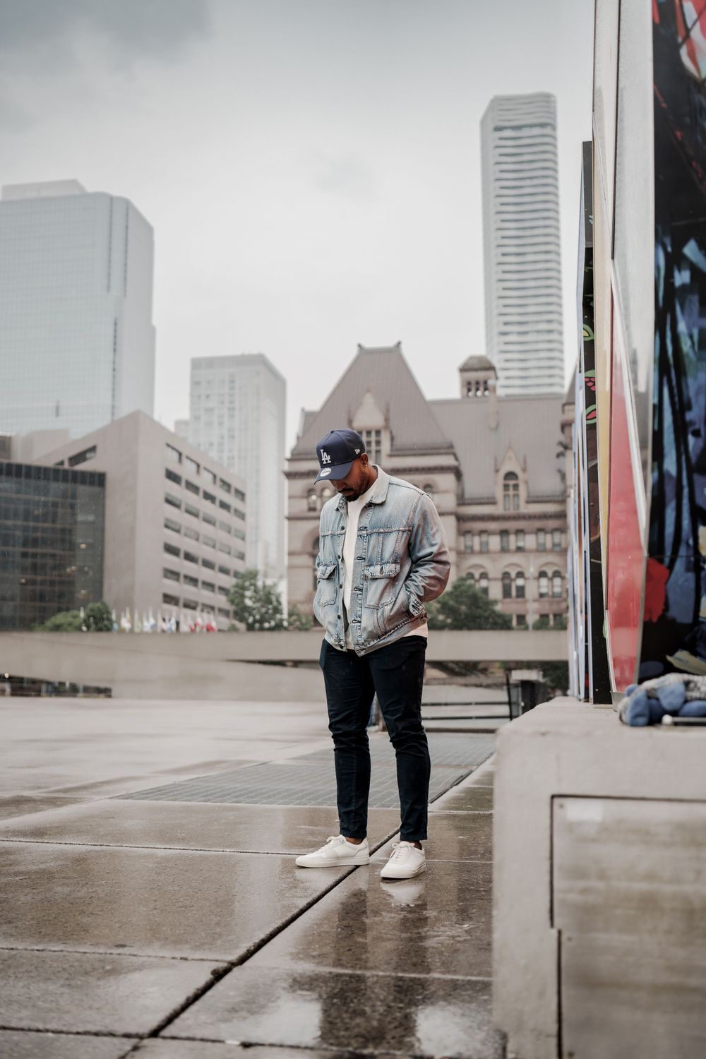A person in denim jacket and baseball cap standing against Toronto cityscape on an overcast day.