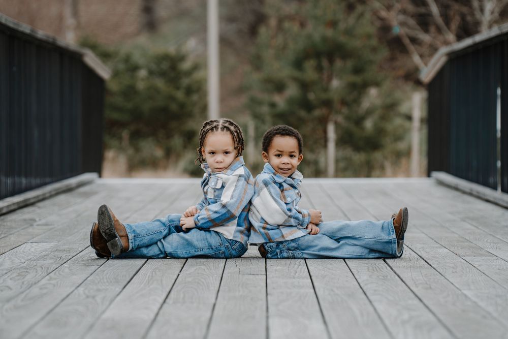 Two toddlers in matching denim outfits sit back-to-back on a wooden bridge.