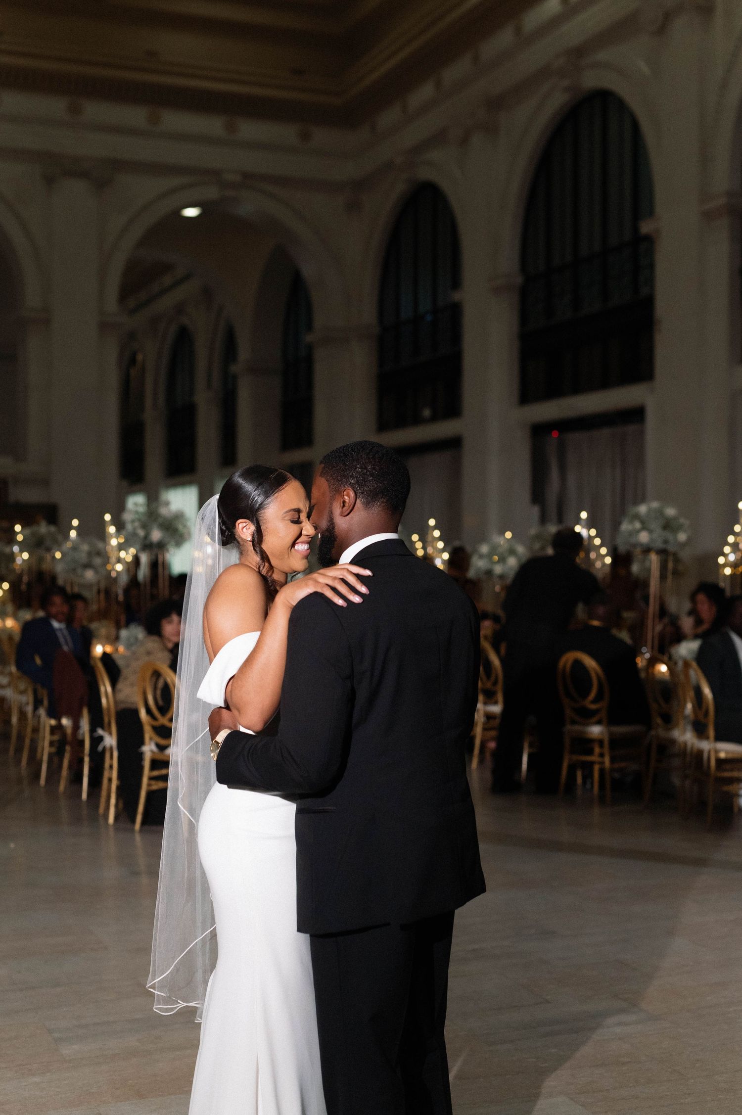 A couple shares their first dance in an elegant ballroom with arched windows and twinkling lights.