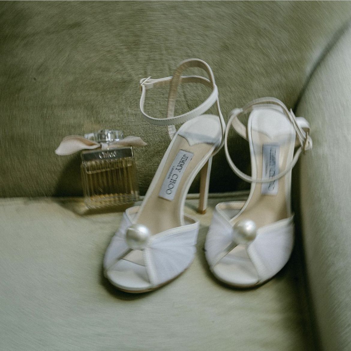 White bridal shoes and perfume bottle arranged on green velvet surface.