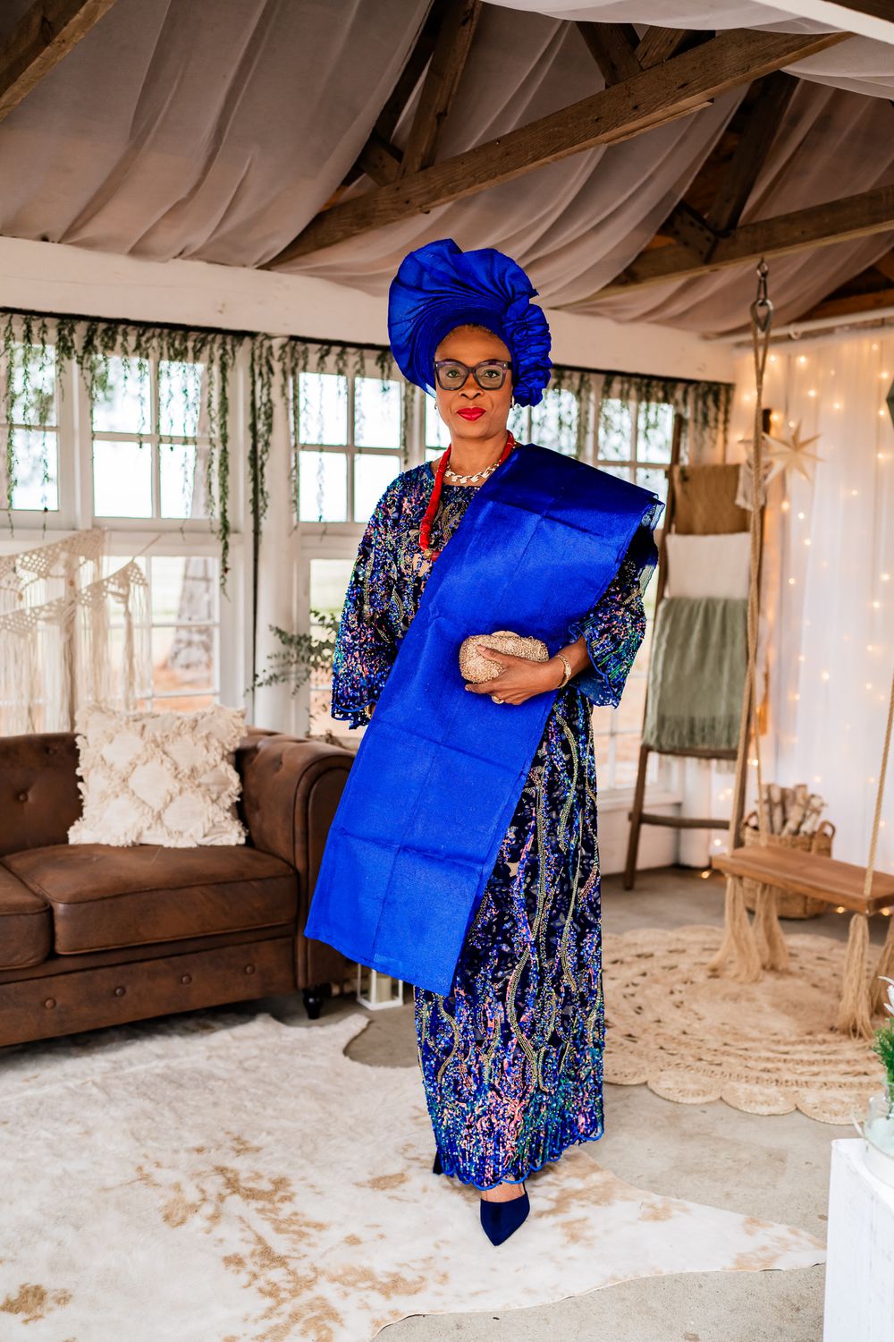 A figure in a vibrant blue dress and matching headwrap poses gracefully in an airy, sunlit room with rustic wooden beams.