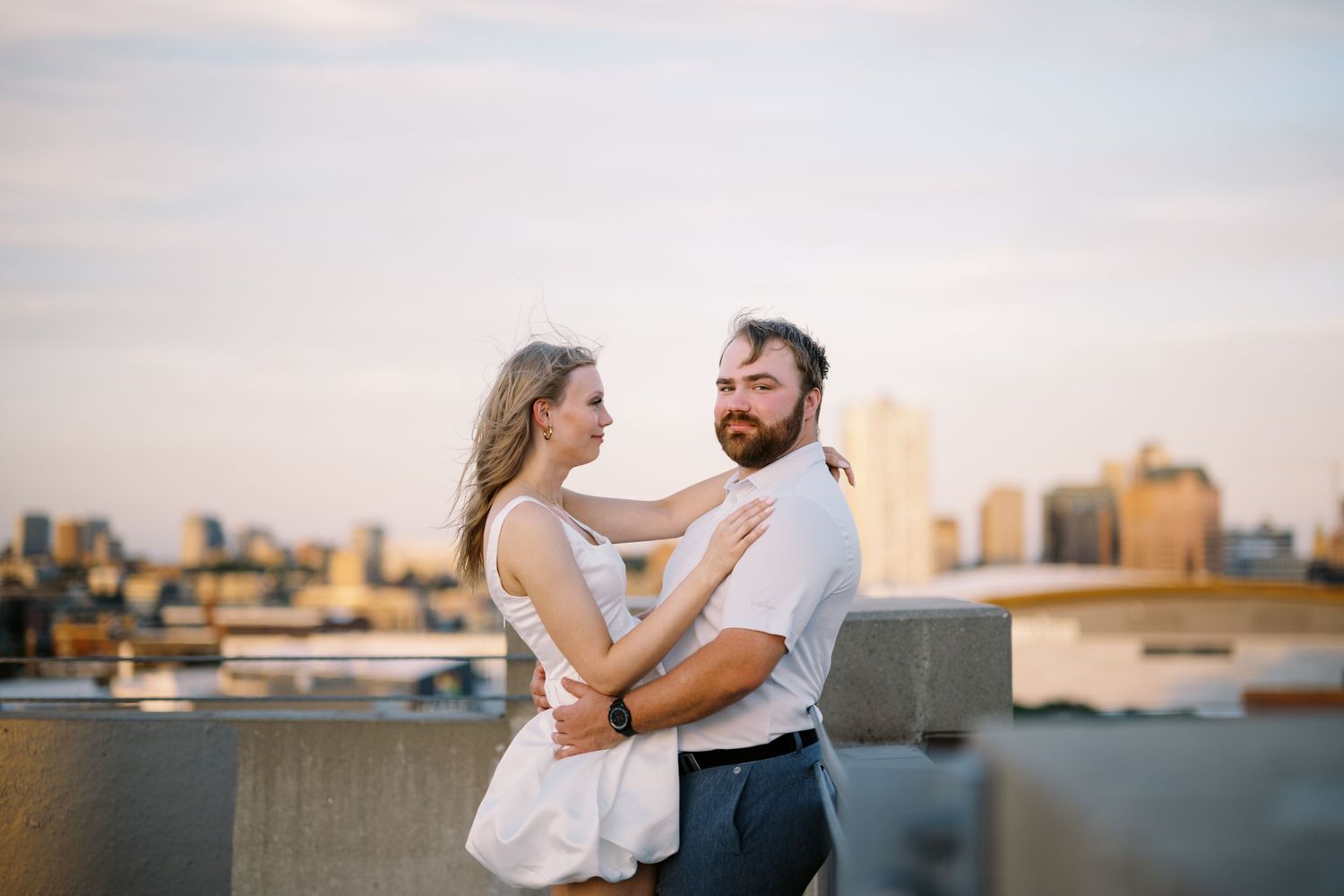 A romantic couple embraces on a rooftop during sunset with a city skyline visible in the background.