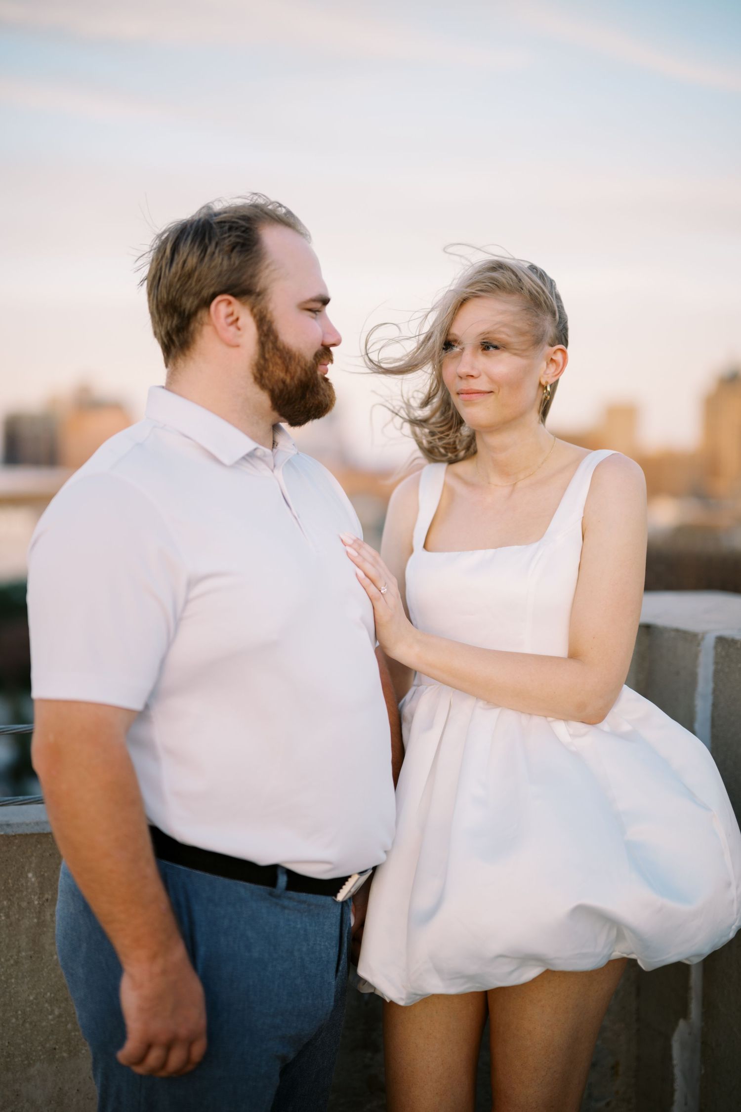 A couple in white outfits shares an intimate moment on a rooftop during sunset with a cityscape background.