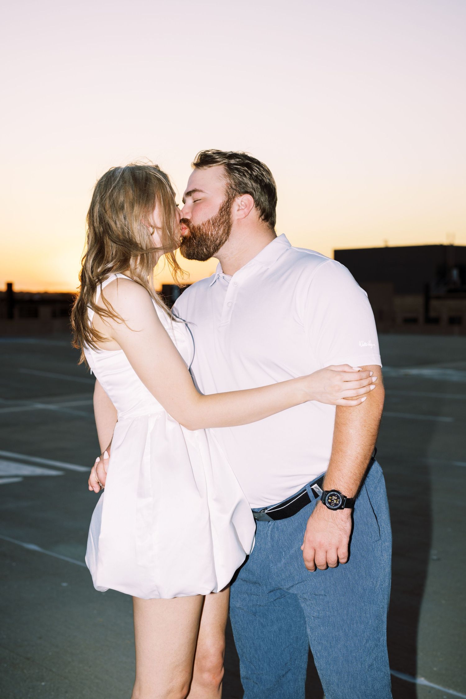 A romantic couple in white and denim share a tender moment at sunset in an empty parking lot.
